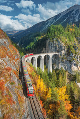 Aerial view of modern red train on Landwasser viaduct in alpine mountains, colorful forest at...