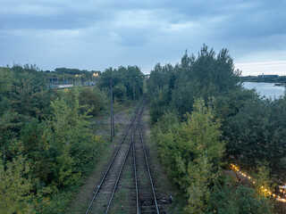 Fototapeta premium Railway tracks through green forest beside river at dusk, elevated view of transport infrastructure and travel logistics landscape