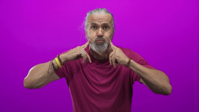 Man with hoary long hair and gray beard points fingers to cheeks in studio with magenta backdrop; playful expression.