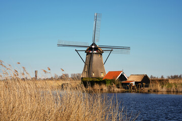 A traditional windmill at Kinderdijk, Netherlands, stands by a calm waterway under a clear blue sky. Surrounded by reeds and historic buildings, it captures the charm of Dutch rural.