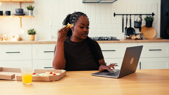 Plus size African American woman types on laptop eating pizza - Powered by Adobe