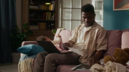 Cinematic shot of young African American father working from home and using laptop while caring for baby, domestic scene lit by sunlight - Powered by Adobe