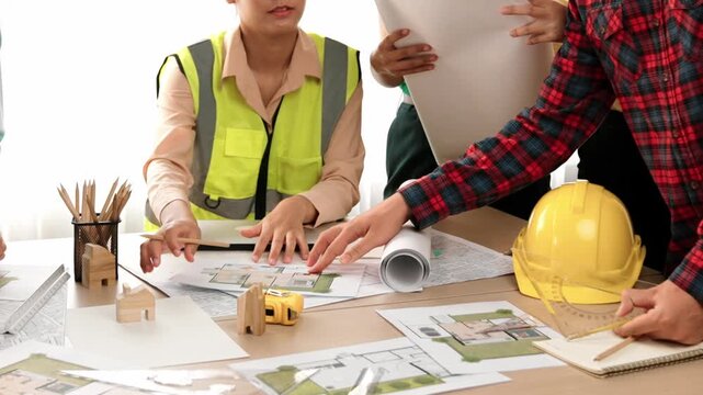 Smart architect and safety engineer discussion about building blueprint on table with architectural document, measurement tools and wooden block scatter around at modern office. Closeup. Delineation.