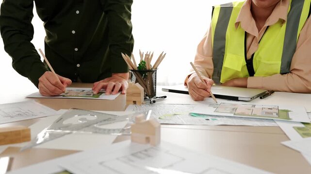 Smart architect and safety engineer discussion about building blueprint on table with architectural document, measurement tools and wooden block scatter around at modern office. Closeup. Delineation.