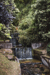 Small cascading waterfall surrounded by lush green trees and stone walls, with clear water gently flowing through a tranquil park setting, creating a peaceful natural retreat