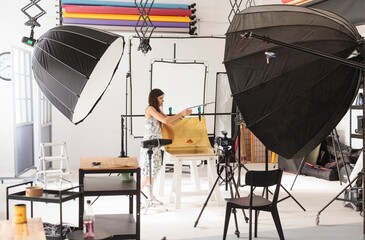In a bright studio, a Woman meticulously adjusts the backdrop and lighting equipment, creating an optimal setting for capturing artful still life