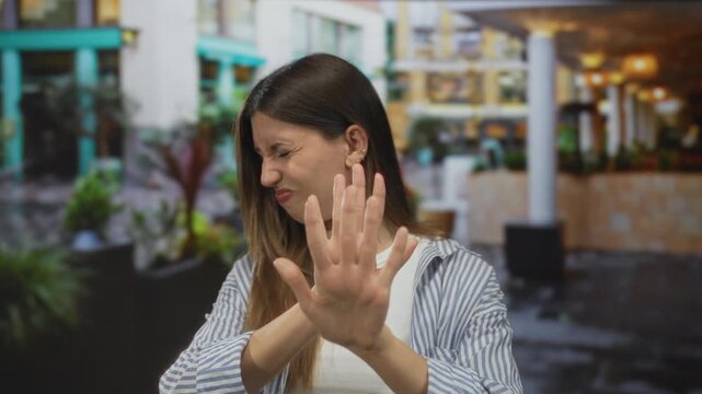Woman holds hand out in stop gesture on city street with a disgusted expression while wearing a striped shirt and white top; rejection.