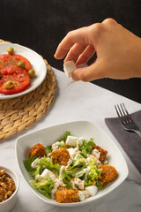 Hand serving diced vegan cheese on a caesar salad with vegan nuggets on a white marble table. A side dish of sliced tomato and a small bowl of nuts are sitting next to it