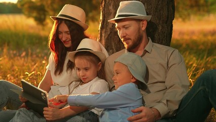 Parents and children play on computer tablet in park under tree. Young family children is sitting on grass in forest relaxing outdoors. Dad mom kids use tablet. Family education with digital gadget