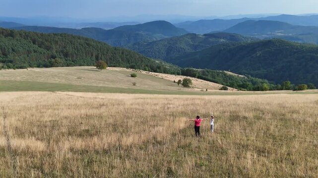A girl and a woman raise their hands and greet a distant passerby as they stand at the foot of a beautiful hill.