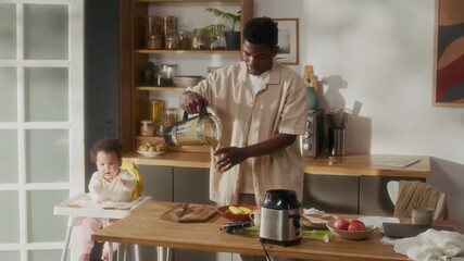 Medium shot of young African American man as father making healthy smoothie while preparing breakfast in kichen with cute baby sitting in high chair, cinematic lighting, copy space - Powered by Adobe