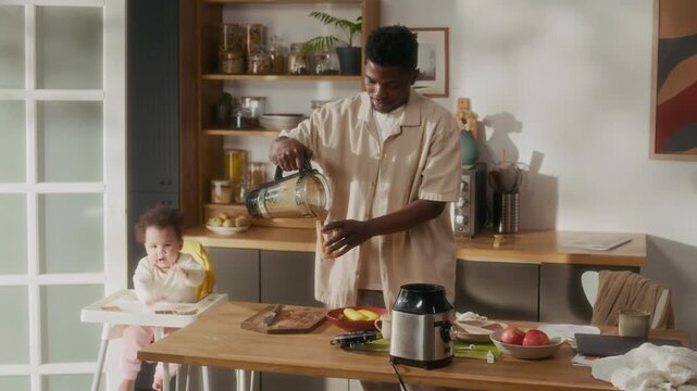 Medium shot of young African American man as father making healthy smoothie while preparing breakfast in kichen with cute baby sitting in high chair, cinematic lighting, copy space