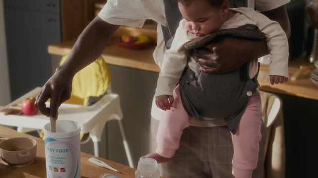 Tilt up shot of young African American man as stay at home father preparing baby formula while holding child in home kitchen