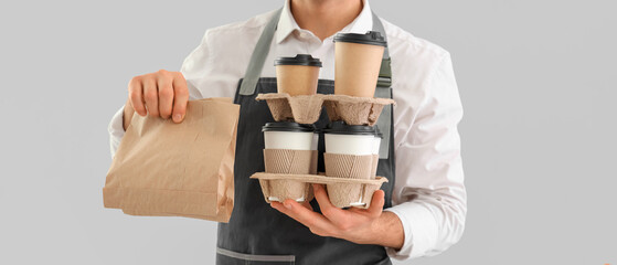 Young male barista with paper cups of coffee and food on grey background, closeup