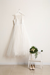 Beautiful wedding dress hanging on white wall, wedding shoes and bridal bouquet in room