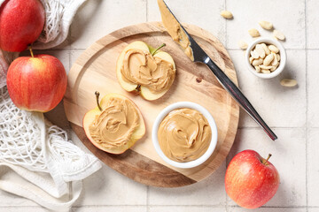 Wooden board with tasty apple slices, nut butter and peanuts on white tile background