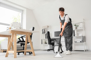 Young male janitor with modern vacuum cleaner cleaning floor in office