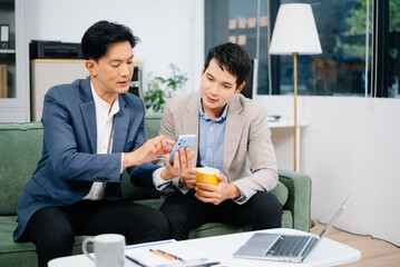 Two Asian businessmen relax with coffee and discuss ideas using a smartphone in a modern office.