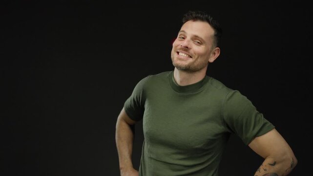Young man smiling confidently against an isolated black background with tattooed arm and green shirt, exuding a handsome and attractive demeanor.
