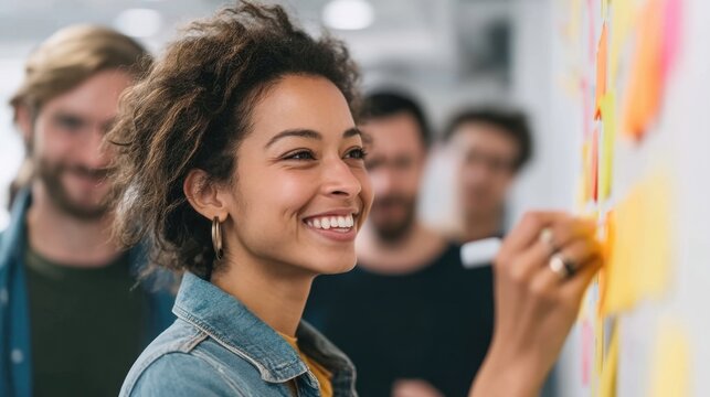 Happy young woman writing ideas on colorful sticky notes in a collaborative workspace with smiling coworkers in the background during a productive team meeting