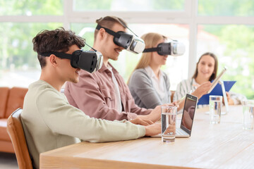 Young man with laptop and his colleagues using VR glasses at table in office