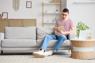 Young man in eyeglasses reading book on sofa at home