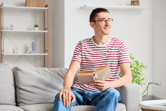 Young man in eyeglasses reading book on sofa at home