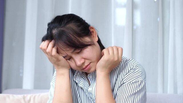 Sad and depressed Asian woman sitting alone, clutching her head in frustration and overthinking life problems, feeling anxious and isolated with stress, headaches, and depression