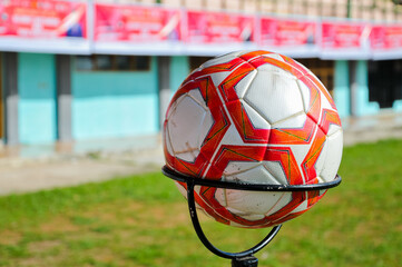 A red and white soccer ball on a holder with a blurred football field in the background, outdoor sports training ground concept.