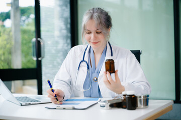 Young female doctor reviewing pill bottle and writing prescription in clinic. Healthcare, patient care, medication review, digital charting. Real people in medical setting.