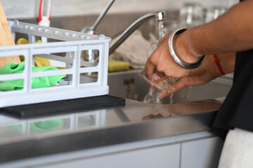 Close up of hands washing glassware under running tap water in a modern kitchen sink with dishes drying rack nearby