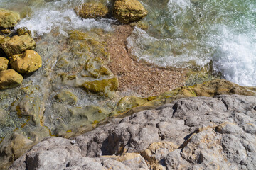 Top-down aerial view of sea waves washing over a textured rocky and pebble shore with clear turquoise water.