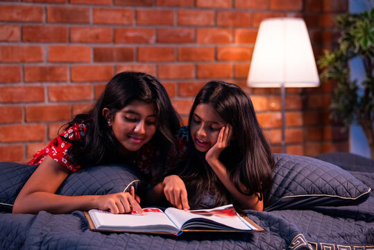 Asian little sisters lying on bed reading a book together indoors with focus and calm expressions