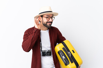 Traveler man man with beard holding a suitcase over isolated white background listening to something by putting hand on the ear