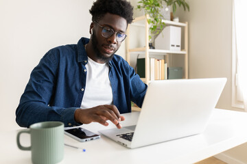 Black man working at home office using laptop and wireless earphone. African American male typing on computer.