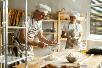Working in bakery - two bakers whisk eggs and sprinkle flour onto dough through sieve