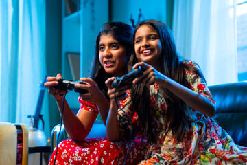 Asian girls playing video game together on sofa with joystick during leisure time at home