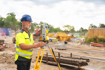 Professional builder  wearing  safety helmet and bright hi-viz clothing uses surveying EDM tool at  busy worksite