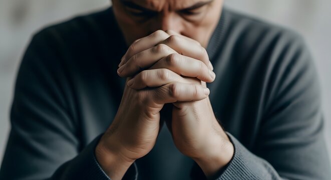 A man clasps his hands together, head bowed in deep thought or prayer, conveying a sense of contemplation and introspection