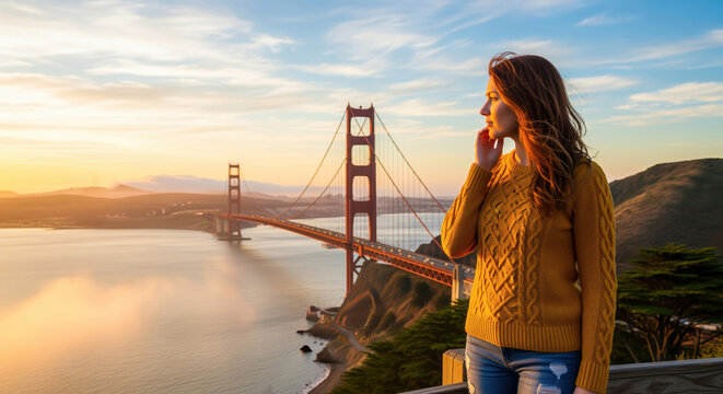 A woman with wind-tossed hair in a golden sweater gazes reflectively at the iconic Golden Gate Bridge during a vibrant San Francisco sunset.