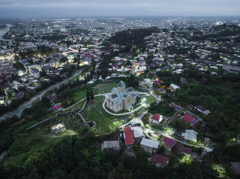 Aerial view of Bagrati Cathedral illuminated against the twilight, nestled among lush greenery, with the cityscape twinkling in the distance, Kutaisi, Imereti, Georgia.