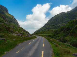 Naklejka premium A winding road cuts through a lush green valley, nestled between imposing mountains under a vibrant blue sky dotted with fluffy white clouds. The scene evokes a sense of adventure and tranquility.