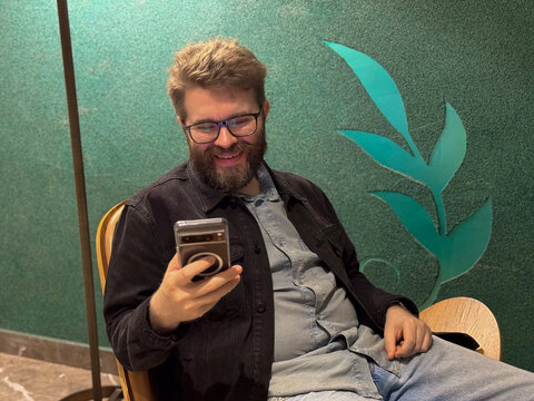 Man smiling while holding a smartphone in a lounge chair. Joyful communication, digital connection, casual lifestyle and relaxation in a modern interior.