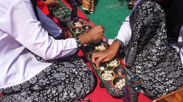 Two people in traditional Javanese attire playing Congklak, a classic Indonesian mancala-style board game with cowrie shells