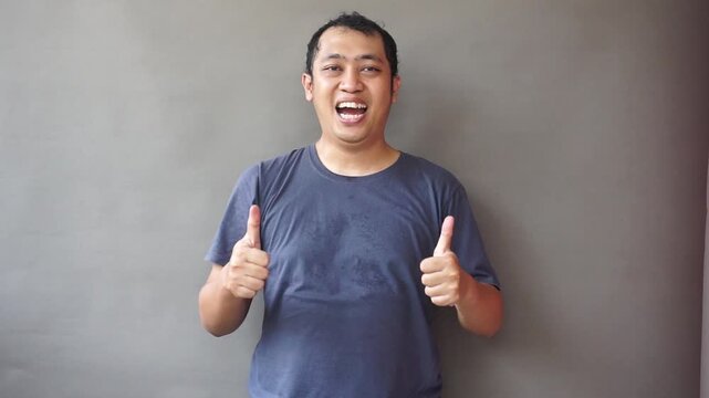 Excited Asian man with an enthusiastic expression gives a double thumbs up gesture for approval and success, standing against a grey background