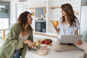Young Women Friends Sharing Morning Conversation During Breakfast Preparation In Contemporary Kitchen With Laptop