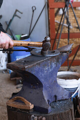 Close-up of blacksmith using hammer and anvil to shape horseshoe, rustic workshop setting with traditional tools and blue barrel in background