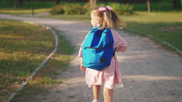 back to school. a little girl with a backpack runs through the green park to school along the path. education kid dream concept. a small child with a backpack back view runs the park sun to school