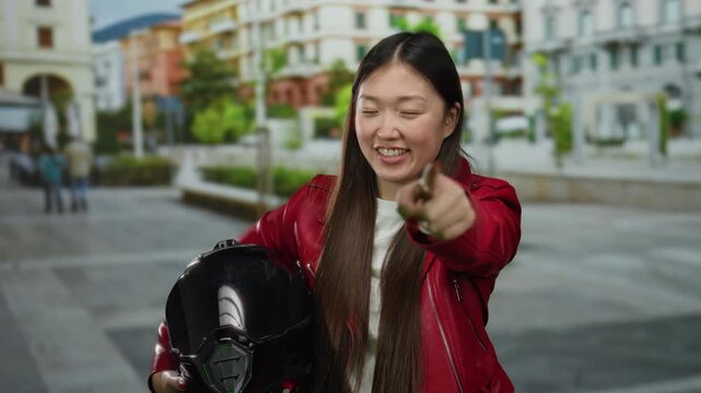Woman wearing red jacket holding motorcycle helmet points on urban street with blurred buildings and people in background.