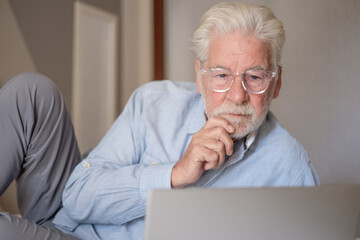 Fototapeta premium Caucasian senior bearded man with eyeglasses lying on bed using computer connectivity browsing internet by laptop enjoying tech and social